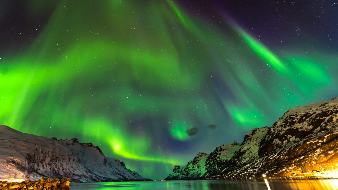 Green aurora over a fjord with the lights of a small village along the shore